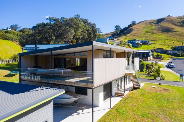 A contemporary multi-level residential home set on a sloping suburban hillside. The building has a low-pitched roof, dark metal framing, light vertically ribbed exterior cladding, expansive glazing, and a large covered upper-level terrace with glass balustrades. The lower level includes an open undercroft or garage-like area, while neighbouring homes, roads, lawns, and rolling hills surround the property.