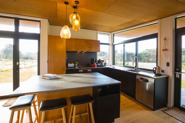 Warm, contemporary kitchen with a large dark stone island and three timber bar stools, black lower cabinetry, timber wall cabinetry, and stainless-steel appliances. Floor-to-ceiling glazing and glazed doors provide expansive views to the surrounding landscape, while pendant lights and a timber-lined ceiling add warmth.