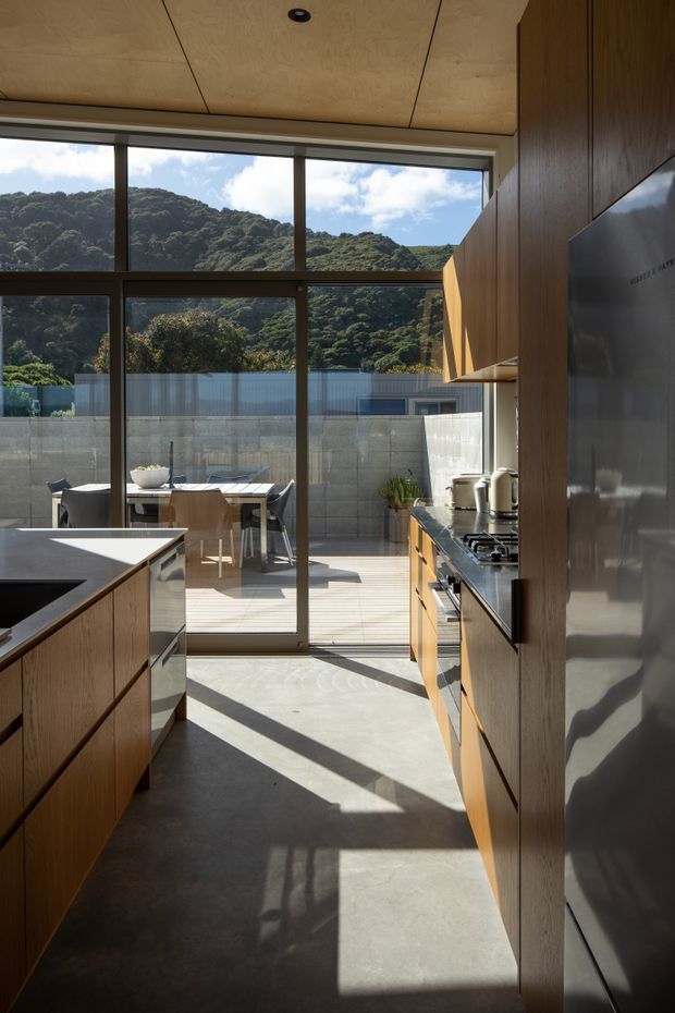 A contemporary kitchen viewed toward a fully glazed outdoor connection. The room features warm timber cabinetry, a central island with a dark benchtop, integrated stainless-steel appliances, and a smooth grey concrete floor. Floor-to-ceiling sliding glass doors open onto a timber deck and outdoor dining area, with a leafy hillside visible beyond. Strong sunlight creates long geometric shadows across the floor.