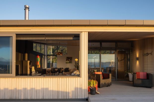 Single-storey modern home exterior with warm vertical timber cladding, broad glazed openings, and a covered outdoor terrace. The image looks toward an open-plan interior dining and living area through the glazing, while the terrace includes outdoor lounge seating and potted plants. A low-profile roof, metal chimney, timber-lined soffit, and strong indoor-outdoor connection are prominent.