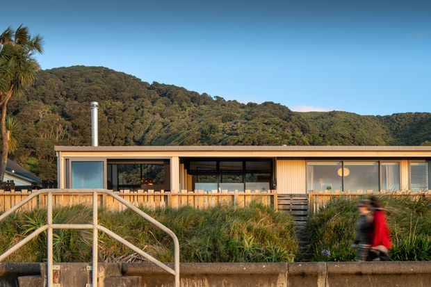Exterior view of a low, single-storey contemporary building set against a densely vegetated hillside. The facade combines warm vertical timber cladding, large dark-framed glazed openings, and a shallow flat or low-pitched roof with a metal chimney. A timber-railed walkway and landscaped grasses occupy the foreground.