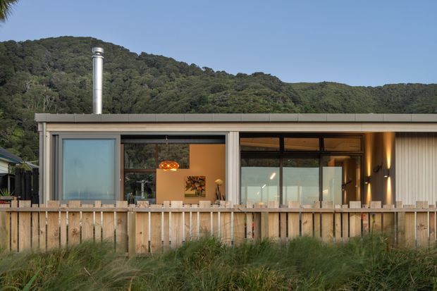 Single-storey contemporary house exterior viewed from a timber deck or front boundary, with extensive floor-to-ceiling glazing, pale vertical cladding, a low-profile roofline, and a prominent stainless-steel chimney. A timber fence and coastal-style grasses occupy the foreground, with densely vegetated hills behind the home.