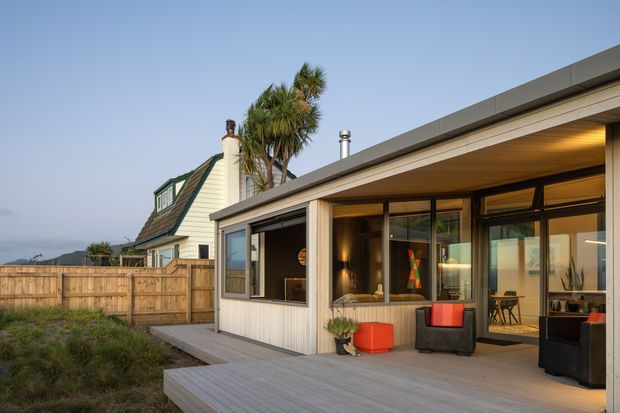 Exterior view of a contemporary single-storey extension with a low-pitched roof, expansive dark-framed glazing, pale vertical wall cladding, and a broad timber deck. The sheltered outdoor living area contains seating and bright orange-red furniture. A separate traditional gabled house and timber boundary fence are visible behind it, with palm trees and distant landscape beyond.