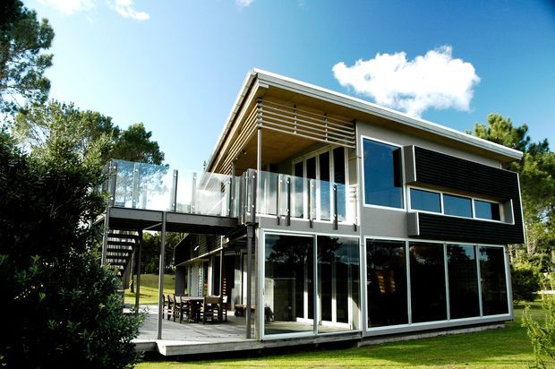 Exterior view of a contemporary two-storey residential house with a low-pitched sloping roof, extensive floor-to-ceiling glazing, dark horizontal cladding, light structural framing, a raised timber deck and glass balustrades. The home is set among mature greenery and lawn, with an external stair and covered outdoor living area visible beneath the upper level.