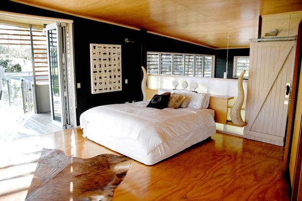 A bright, compact bedroom with a low timber-lined ceiling and polished timber flooring. The room features a white bed with layered pillows, a black feature wall with framed artwork, louvred windows, a white built-in headboard and bedside storage, and a tall white barn-style wardrobe door. Bi-fold glazing opens to a timber outdoor deck, bringing in abundant natural light. A cowhide rug adds a rustic accent.