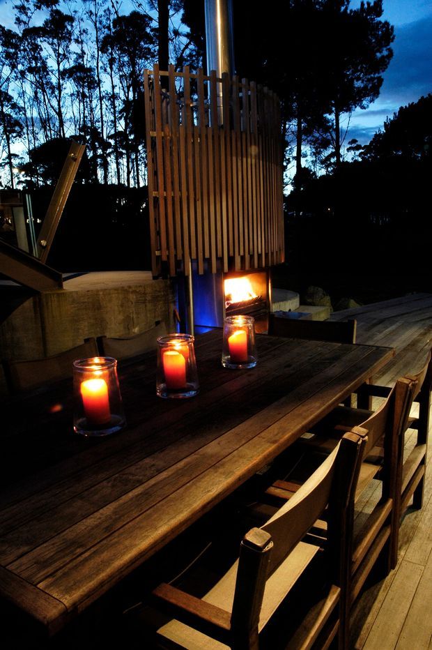 A moody outdoor dining area at dusk, centred on a long dark timber dining table with timber chairs and several glowing candles. A sculptural outdoor fireplace or fire feature with vertical timber screening rises behind the table, set on a timber deck surrounded by silhouetted trees.