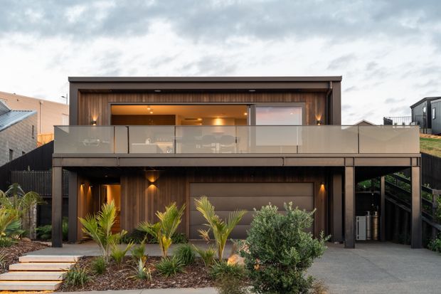 Front exterior view of a contemporary two-storey residence with a dark timber-clad facade, broad upper-level glazing, a frosted glass balcony balustrade, covered lower-level parking or entry space, and landscaped planting along the driveway.