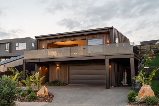 Contemporary two-storey residential façade with dark vertical timber cladding, a broad upper-level glazed balcony, recessed warm lighting, and an integrated double garage. The home is set on a landscaped sloping suburban site with low-maintenance tropical planting and feature rocks.