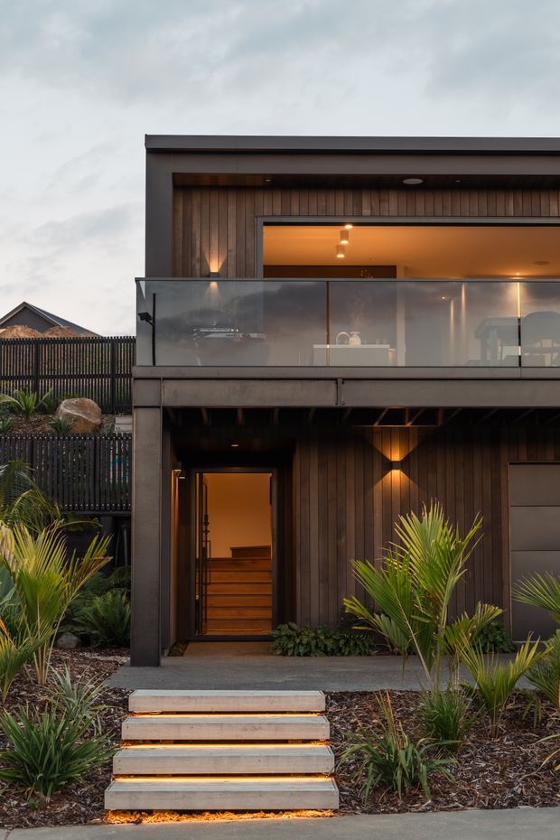 Front exterior view of a contemporary two-storey residence with dark vertical timber cladding, a projecting upper balcony with translucent glass balustrade, charcoal metal framing, and a recessed illuminated entry. Concrete steps with integrated warm lighting lead through a landscaped garden of ornamental grasses and palms.
