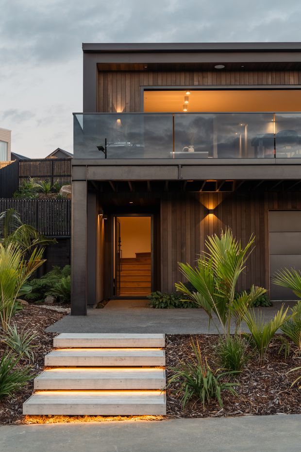 Front exterior view of a contemporary multi-level residence at dusk, featuring dark vertical timber cladding, a broad upper balcony with clear glass balustrading, recessed warm lighting, a sheltered entry, and illuminated concrete steps set among landscaped tropical planting.