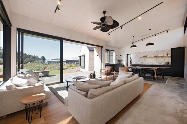 Open-plan contemporary living area with a large cream sectional sofa and armchairs arranged around low coffee tables, opening directly to a kitchen and dining zone. Floor-to-ceiling black-framed glazing provides views to the surrounding landscape. The space features a polished concrete floor, warm timber flooring in the seating area, a dark kitchen island with bar seating, a marble-look splashback, track lighting, and a ceiling fan.