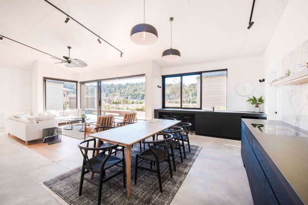 Bright open-plan kitchen, dining, and living area with large black-framed windows and sliding doors overlooking a leafy landscape. A long timber dining table with black chairs sits on a textured rug, while a white sofa occupies the adjacent living zone. The kitchen features dark fixed cabinetry, pale benchtops, open shelving, and minimalist black track and pendant lighting. The space has a clean contemporary aesthetic with abundant natural light.
