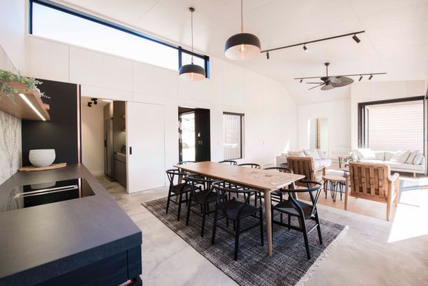 Bright open-plan interior combining a kitchen, dining area, and living room. The space has white walls and ceiling, polished concrete flooring, a dark kitchen island with integrated sink and cooktop, a long timber dining table with black chairs, and a light lounge area with generous windows. Black pendant lights, track lighting, and a ceiling fan add contrast. High-level glazing and large windows create an airy, contemporary feel.