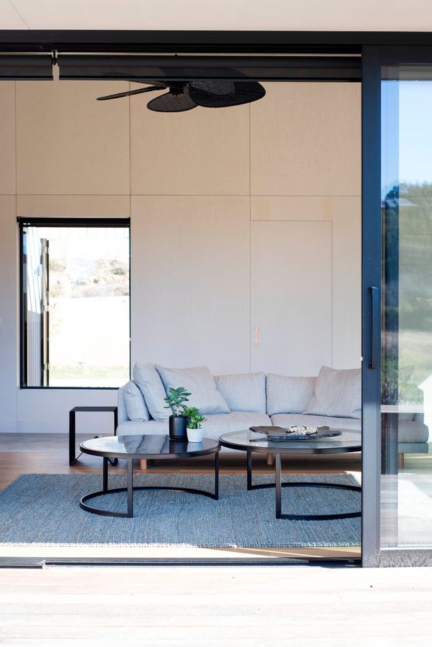 A bright, minimalist living room viewed through a large open sliding glass door. The room features a pale sectional sofa, two round black-framed coffee tables, a blue-grey area rug, pale wood flooring, light paneled walls, dark ceiling-mounted curtain or blind tracks, and a black ceiling fan. Large glazed openings connect the room to the exterior.