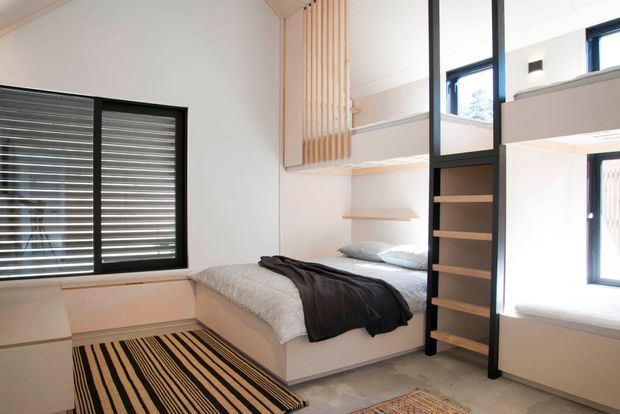 Bright compact bedroom with a built-in platform bed, pale upholstered or painted joinery, open shelving, and a black-framed internal partition or ladder-like storage element. Black-framed windows with horizontal blinds provide daylight. Vertical timber battens form a decorative feature near the upper wall, while striped rugs and dark bedding add contrast. The room has a clean, contemporary built-in design with a loft-like upper-level opening visible behind the partition.