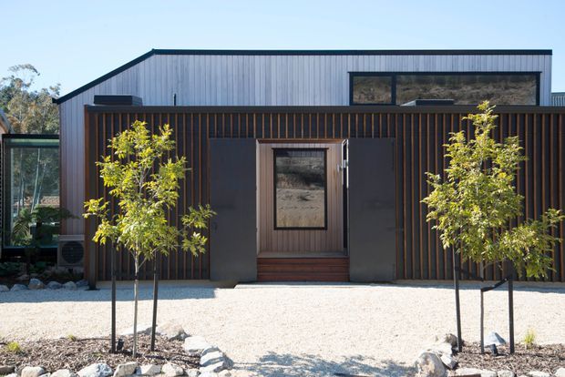 A modern single-storey residential facade with a flat roof and a layered composition of pale vertical metal cladding above dark vertically slatted timber or wood-toned screening. A recessed central entry door is framed by dark cladding, with narrow full-height glazing at the sides and a gravel forecourt landscaped with young trees and rocks.