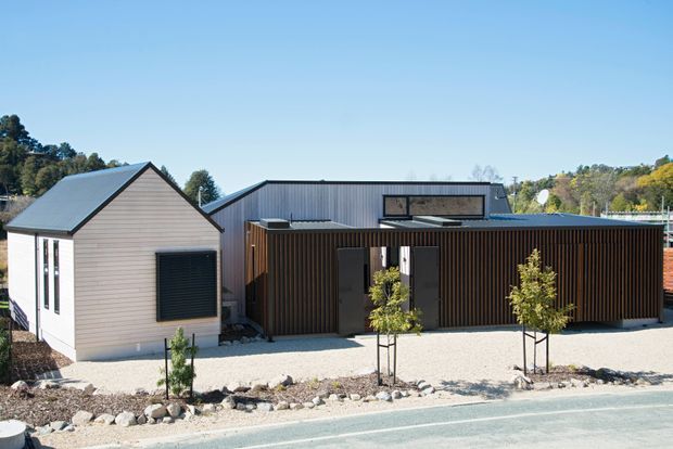 A contemporary single-storey building viewed from the street, combining a gabled light-coloured weatherboard volume with a larger flat-roofed metal-clad section and a projecting dark vertical timber-screened entry or outdoor area. The landscaped frontage uses pale gravel, rocks, young trees and restrained planting. Clear blue sky and surrounding mature vegetation frame the building.