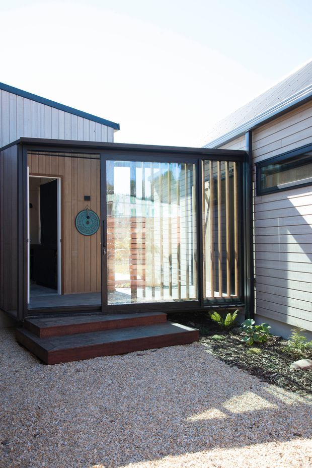 Exterior view of a contemporary single-storey home centred on a glazed entry link or enclosed courtyard connection. The facade combines pale horizontal and vertical timber-style cladding with dark metal-framed floor-to-ceiling glazing, a low sloping metal roof, and broad timber steps leading from a gravel forecourt. An interior doorway and planted garden bed are visible around the entry.