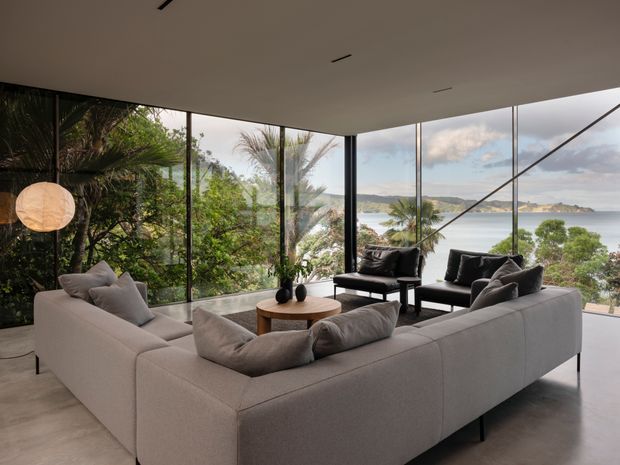 A refined contemporary living room arranged around a low round coffee table, with a large pale sectional sofa and two dark lounge chairs. Floor-to-ceiling corner glazing opens to expansive coastal and waterfront views, while lush palms and greenery frame the interior. The room has a minimalist flat ceiling, slim dark window frames, concrete-toned flooring, and soft neutral furnishings.