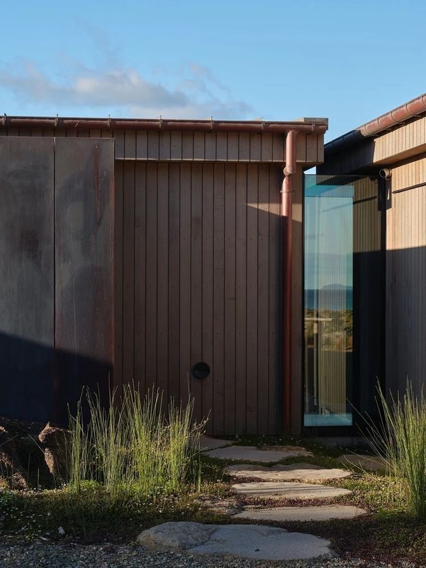Exterior view of a compact contemporary structure with dark vertical timber cladding, weathered metal wall panels, exposed copper-toned rainwater pipes and a narrow full-height glazed doorway. Irregular stone stepping slabs lead through a naturalistic grass landscape, while the glazing reflects a distant coastal or landscape view. The low roofline and simple material palette create a restrained, earthy modern appearance.