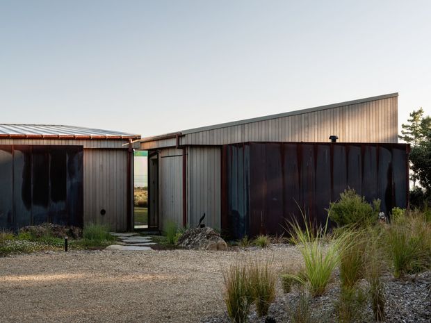 Single-storey contemporary building exterior composed of connected shed-like volumes, with vertical corrugated metal cladding, dark metal doors or screens, and a low-pitched asymmetrical roofline. A gravel forecourt and naturalistic grasses surround the entrance, while a narrow glazed opening provides a glimpse through the building.