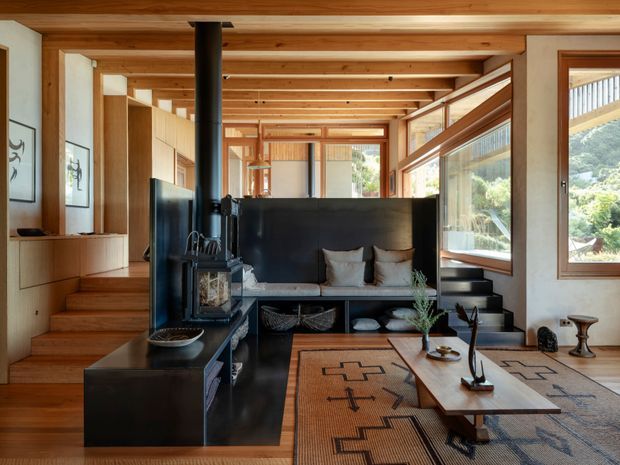 Warm contemporary living room with a compact black wood-burning stove, built-in timber cabinetry, stepped seating, exposed timber ceiling beams, large windows, and a low coffee table over patterned rugs. The room is connected to an open circulation area and appears to include a split-level stair arrangement.
