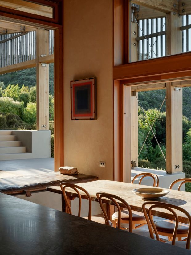 A warm, light-filled open-plan dining and kitchen area with a timber dining table and curved wooden chairs in the foreground. The room features large timber-framed openings and glazing, exposed timber posts and beams, warm earthen plaster walls, and views toward a lush wooded landscape. A raised concrete seating or platform area is visible beside the dining space, with a dark kitchen benchtop occupying the lower foreground.