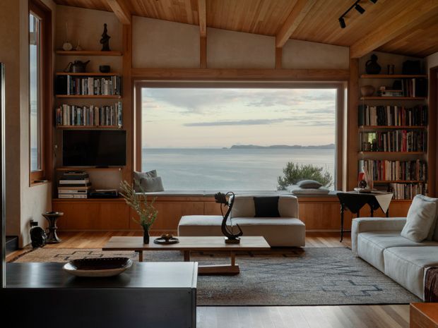 Warm timber-lined living room with built-in bookshelves, a pitched timber ceiling, a large picture window overlooking the sea, and contemporary sofas arranged around a low coffee table. The room has a refined coastal outlook with dark and natural wood accents.