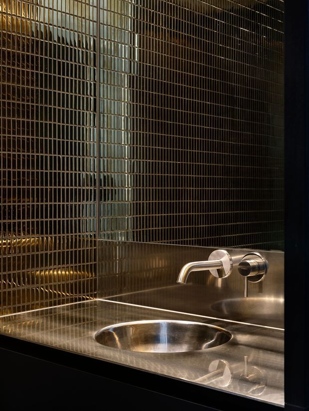 Close-up of a refined bathroom vanity featuring a round integrated stainless-steel basin, wall-mounted chrome-silver tapware, and a reflective metallic benchtop and backsplash. The walls are covered in small vertically ridged metallic tiles, creating a dark, lustrous and highly reflective finish.