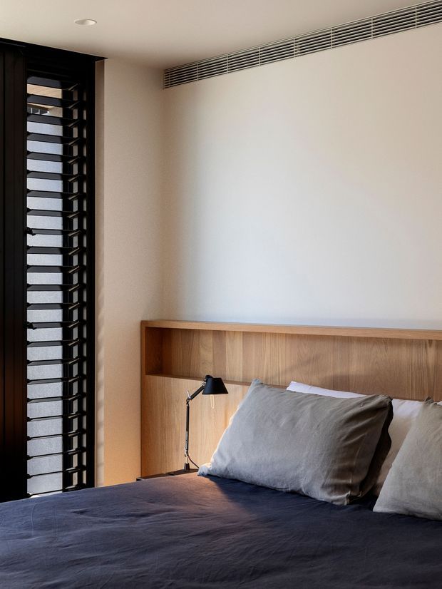 A calm, contemporary bedroom corner with a built-in timber bedhead and integrated bedside shelf, layered with neutral grey bedding and pillows. A black reading lamp sits beside the bed, while a tall black-framed louvred window brings in filtered light. A linear ceiling ventilation grille spans the upper wall.