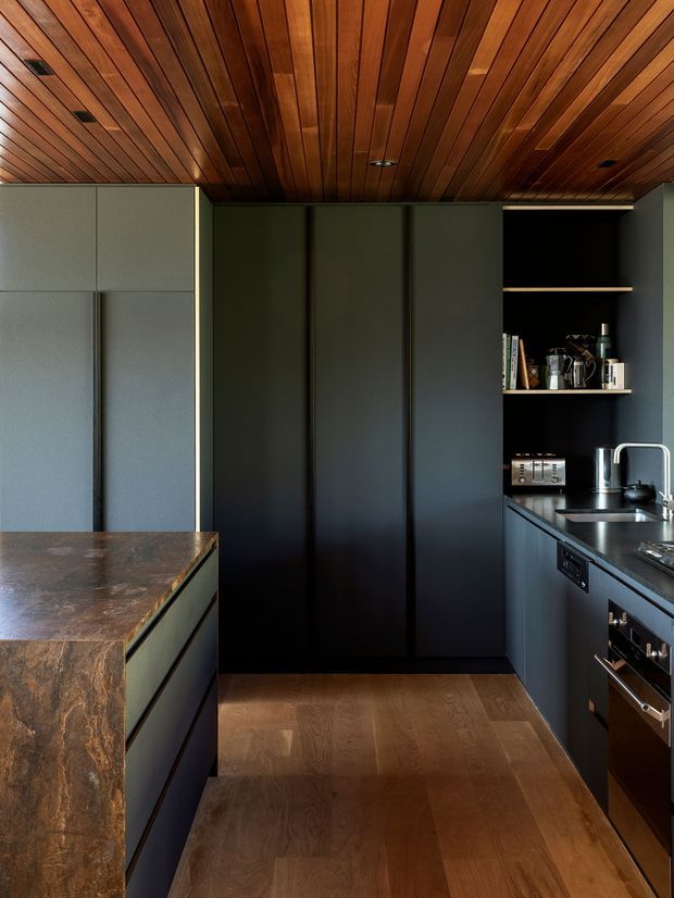 Compact, sophisticated galley kitchen with dark matte cabinetry, integrated appliances, open shelving, and a richly veined stone island benchtop. Warm timber boards line the ceiling and continue as the flooring, creating a strong contrast with the charcoal joinery. Stainless steel sink and tapware sit beneath open shelves displaying kitchenware, while recessed ceiling lights provide discreet illumination.