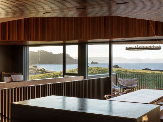 A warm, timber-lined open-plan kitchen and living space with a large central island and dining table, overlooking a rugged coastal inlet through expansive black-framed windows. Vertical timber screening, a timber slatted ceiling, dark cabinetry, and an outdoor balcony with lounge seating create a refined connection to the landscape.