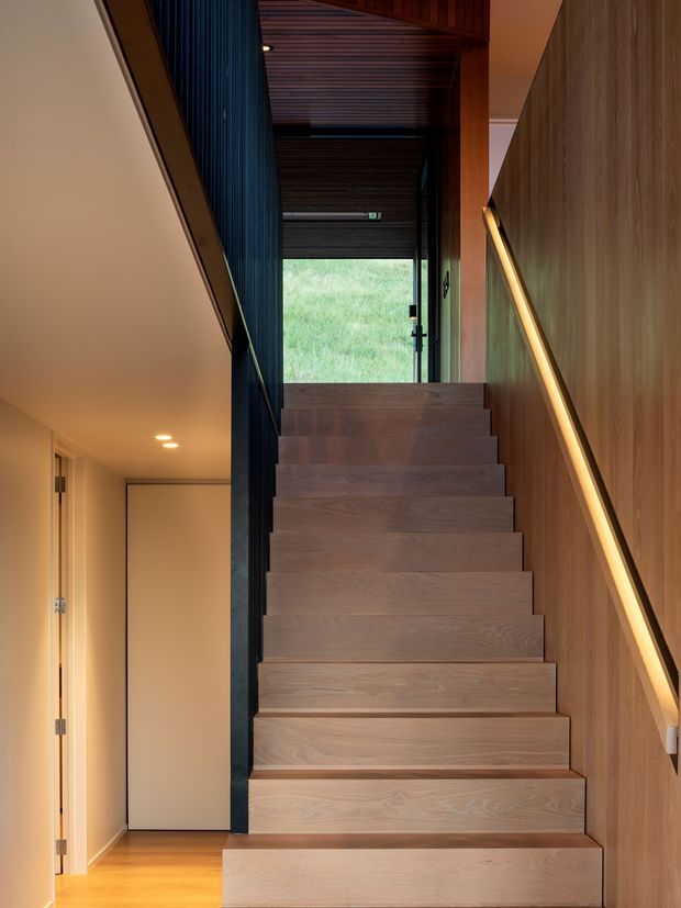 Interior view looking up a straight timber staircase in a narrow circulation space. The stair treads and risers, adjacent wall panelling, and slatted ceiling are warm natural wood, contrasted with a dark vertical metal or timber screen. A slim integrated illuminated handrail runs along the right wall, while a glazed door or window at the top opens toward a grassy exterior. A simple hallway is visible to the left.