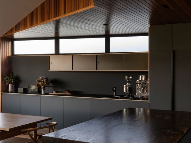 A contemporary kitchen with a long run of dark matte cabinetry, a black benchtop, open shelving, and integrated appliances. A large dark stone or textured island dominates the foreground, with a timber dining table and chairs at left. High horizontal clerestory windows provide natural light, while the ceiling features warm timber slats and a contrasting light-toned panelled section. Coffee equipment and decorative vessels sit along the rear counter.