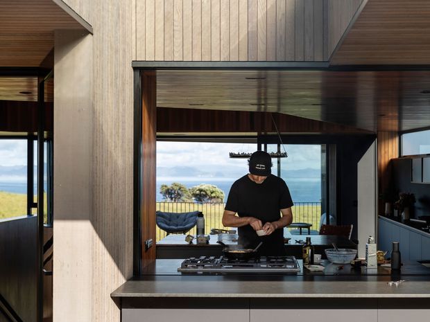 A contemporary open-plan kitchen viewed from behind a dark stone island bench. A person is cooking at a built-in gas cooktop, with timber-lined ceilings and walls, dark window and door frames, integrated cabinetry, and expansive glazing opening toward a coastal ocean view and outdoor seating. The space has strong indoor–outdoor connection and warm natural timber detailing.