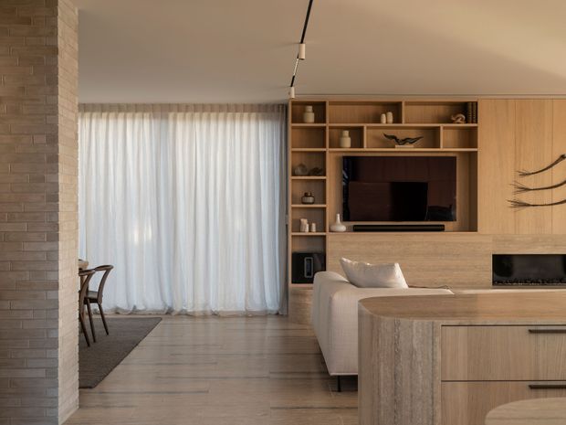 Warm contemporary open-plan living area with a pale timber built-in entertainment wall, integrated shelving and television, a rounded timber kitchen island in the foreground, and a light upholstered sofa. A pale brick column frames the left side, while sheer curtains provide soft daylight and light timber flooring continues through the space. Minimal black track lighting and subtle sculptural wall decor add restrained detail.