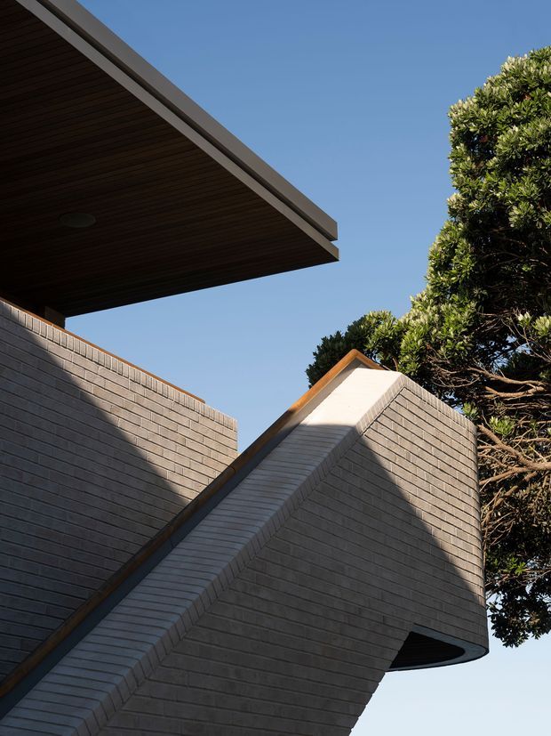 Close-up exterior architectural view of a contemporary building composed of dark grey brick walls, a projecting flat roof with a dark timber-lined soffit, and a sharply angled gabled volume. A rounded projecting brick form and mature planting frame the composition against a clear blue sky.