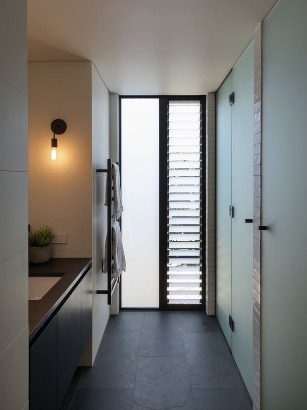 A narrow, contemporary bathroom with a dark tiled floor, pale large-format wall tiles, dark built-in vanity cabinetry and a black benchtop. A frameless glass shower enclosure occupies the right side, while a tall frosted window and black louvred window provide filtered natural light. A wall-mounted black pendant light, towel rail, and small potted plant add detail.