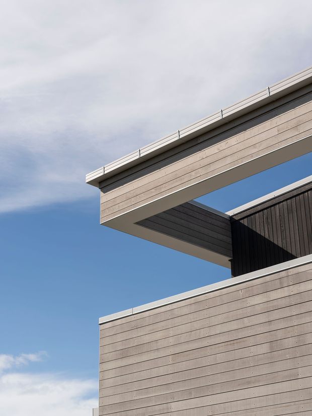 Upward-looking close-up of a contemporary building exterior with layered horizontal timber-clad volumes, a deep cantilevered roof or upper floor, and dark vertical cladding recessed beneath it. The facade has crisp geometric lines and light grey-brown weathered timber tones against a clear blue sky.