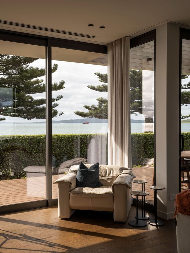 A calm, light-filled living room arranged around a tan leather armchair, with full-height glazed sliding doors opening to a timber deck and waterfront outlook. Sheer curtains, dark window frames, timber flooring, side tables, and a hedge-lined exterior contribute to a refined coastal atmosphere.
