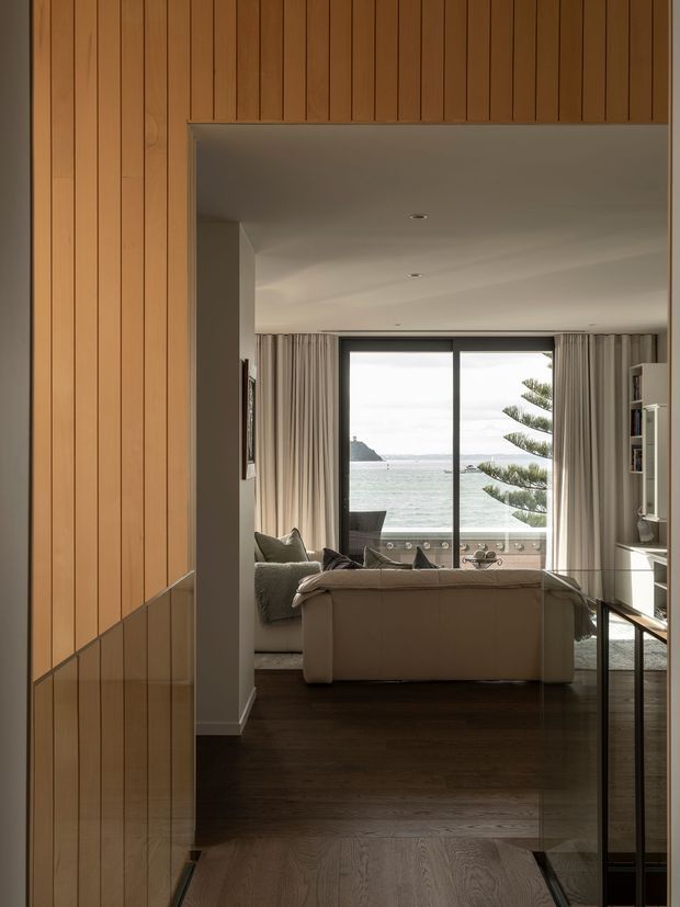 View from a timber-lined hallway or entry toward a refined living room. Vertical warm timber wall panelling and a glazed stair balustrade frame the space, while the living room has dark timber flooring, a pale sofa, curtains, built-in shelving, and large sliding glass doors opening to a balcony with a clear coastal or waterfront outlook.