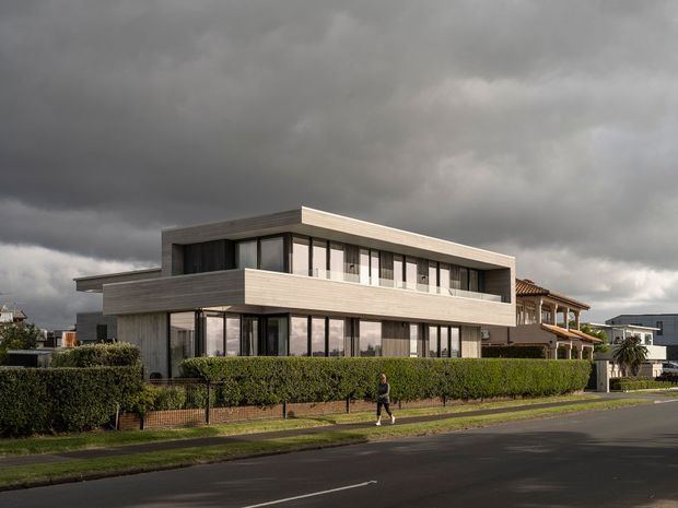 Exterior photograph of a contemporary multi-level residence on a suburban street. The house has strong horizontal massing, extensive dark-framed glazing, projecting upper-floor volumes, and light timber-toned horizontal cladding. A clipped hedge and brick boundary wall frame the front garden, with neighbouring homes visible under a dramatic overcast sky.