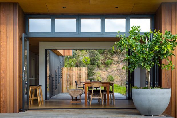 A warm, modern indoor-outdoor dining space framed by extensive timber-clad walls and ceilings. Full-height sliding glass doors open onto a covered terrace or courtyard, with a timber dining table and chairs, bar stools, and a large potted citrus tree. High clerestory windows bring additional light into the timber-lined room, while a rugged planted hillside is visible beyond.