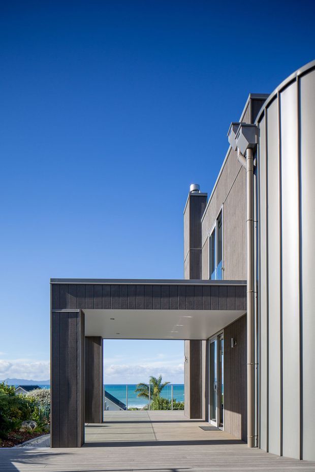 Exterior architectural view of a contemporary coastal residence, featuring a dark timber-clad entry structure framing a covered passage toward ocean views. The building combines vertical dark cladding, pale curved metal wall panels, large glazed openings, and a broad timber deck under a clear blue sky.
