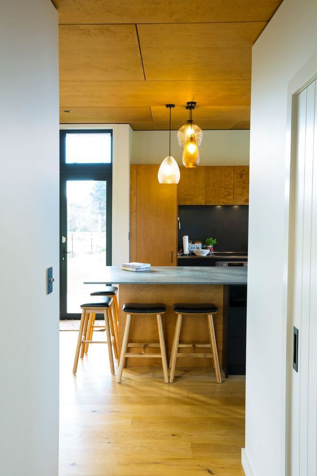 A warm, compact contemporary kitchen viewed through a doorway, featuring extensive timber cabinetry and ceiling panelling, a dark backsplash, a long pale stone island with timber-and-black bar stools, pendant lighting, timber flooring, and a glazed exterior door.