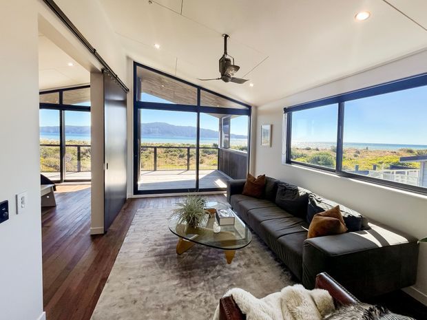 Bright contemporary living room with a large dark sectional sofa, glass coffee table, pale area rug, and ceiling fan. Full-height sliding glass doors open to a balcony or terrace, while expansive windows frame a coastal landscape and ocean view. Dark timber flooring continues into the adjacent space, with white walls and ceiling creating an airy interior.