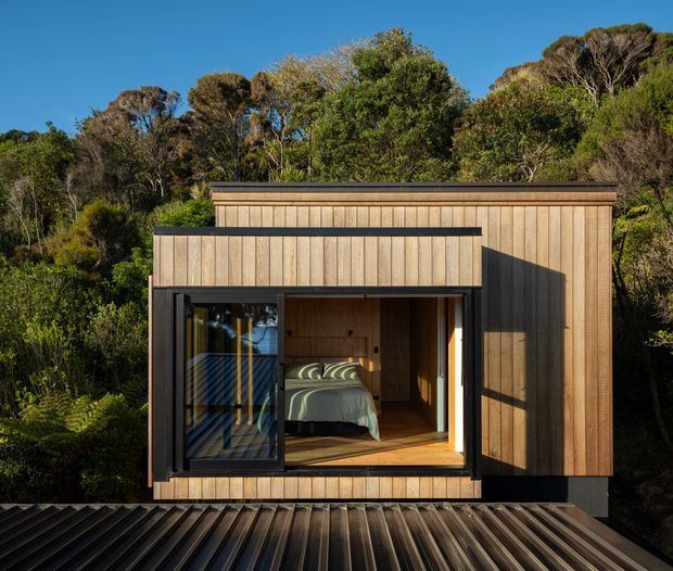 Compact timber-clad cabin set within dense native vegetation, photographed from outside. The simple rectangular form has a low flat roof, dark metal trim, and a large glazed opening revealing a small bedroom with a bed and timber interior. A corrugated metal roof occupies the foreground.