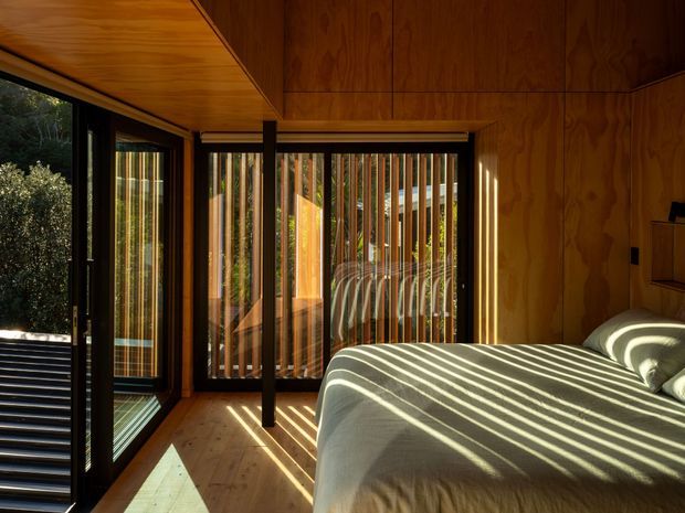 A compact, warmly lit bedroom enclosed in rich timber panelling, with a timber-lined ceiling and timber floor. Full-height black-framed glazing and doors open toward greenery, while vertical external screening casts strong striped sunlight across the bed and floor. The room has a calm, natural, cabin-like atmosphere and a strong indoor–outdoor connection.