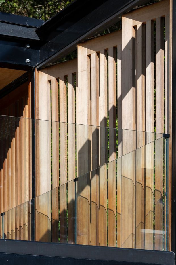 Close-up architectural detail of an exterior balcony or terrace balustrade. Vertical pale timber slats form a privacy screen, supported by a dark metal roof and frame structure, with clear glass panels and dark metal edging in the foreground. Strong sunlight creates geometric shadows across the timber.
