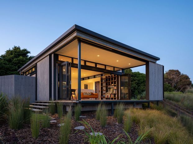 A compact contemporary single-storey pavilion-style home set within landscaped grasses. The dark vertical-clad structure has a broad flat roof with deep overhangs, extensive corner glazing and open bi-fold or sliding doors. Warm interior lighting reveals a living area with timber shelving and built-in joinery, while a raised timber deck and steps connect the house to the planted garden.